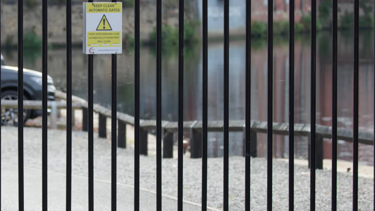 A black metal security gate swings open beside a brick wall, revealing a riverside path in daylight. Camera remains steady, natural lighting
