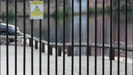 a black metal security gate swings open beside a brick wall, revealing a riverside path in daylight. camera remains steady, natural lighting