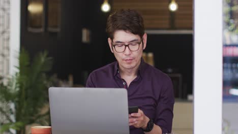 Asian-man-with-laptop-talking-on-smartphone-while-sitting-at-a-cafe