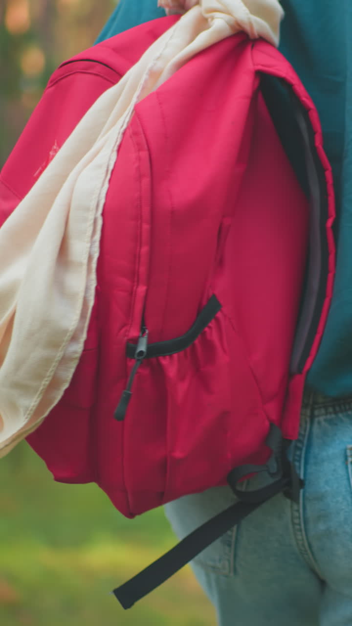 Back view of woman walking through sunlit forest with red backpack slung over her shoulder, adjusting bag with one hand, she wears a green shirt, blending with the natural surroundings
