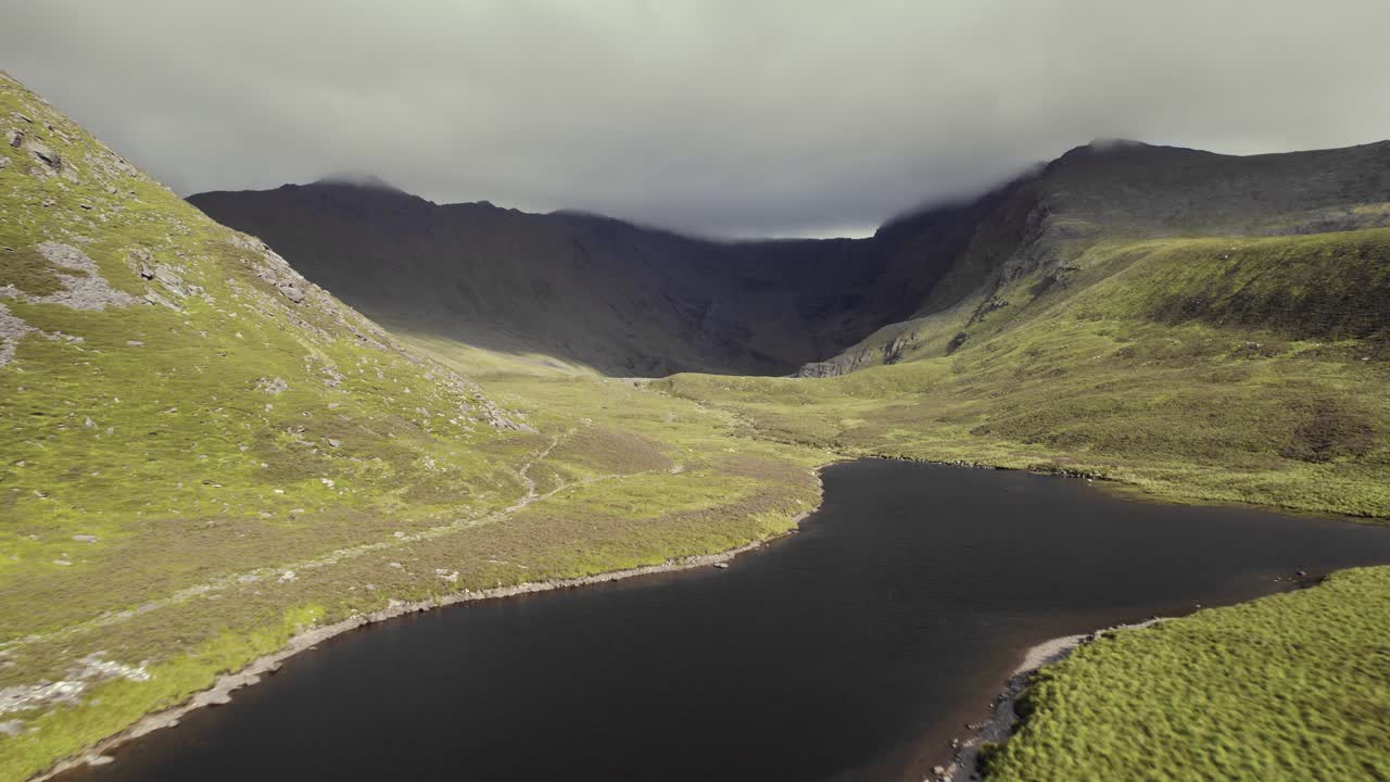 hermosa antena de un pequeño río con un lago dentro de montañas cubiertas de hierba con una cordillera cubierta de nubes de piedra que se encuentra detrás