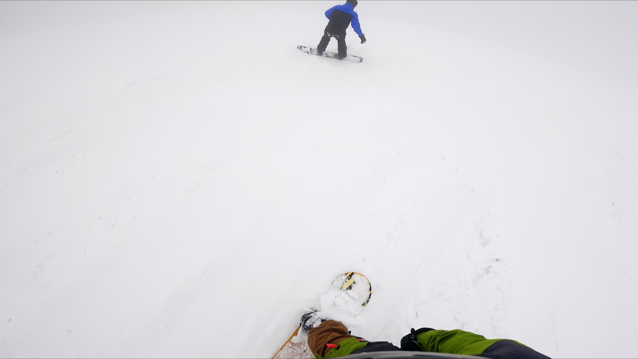 One snowboarder is catching up with his fellow descending from the mountain slope. First person footage of a snow ride.