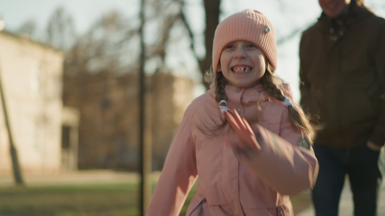 A cheerful little girl in a pink cap and jacket runs with a big smile, while a man in a brown jacket and blue jeans follows behind her