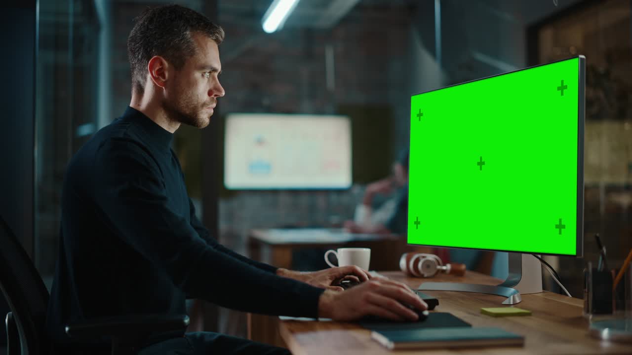 giovane e bello specialista che lavora su un computer desktop con schermo verde in un ufficio creativo affollato con i colleghi. il manager maschio con la barba tagliata indossa un maglione nero casuale.