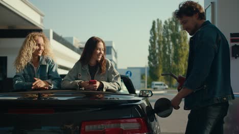 A-group-of-happy-friends-and-a-gray-convertible:-two-girls-watch-a-guy-fill-up-his-car.-Happy-car-ride,-gas-station-stop