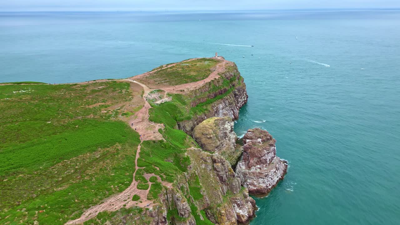 Drone approaching Cap Fréhel lighthouse with coastal cliffs, trails, and the sea in view.