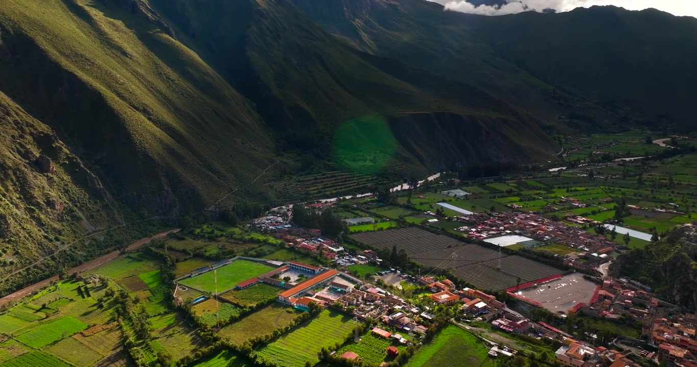 Vista con drone di un insediamento storico annidato in una profonda valle, circondato da montagne ripide e campi verdi a Ollantaytambo, Perù.