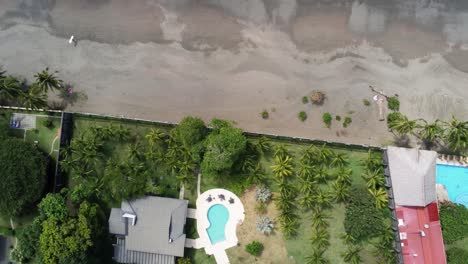 Luftaufnahme-Von-Oben-Nach-Unten-Tropischer-Strand-Mit-Hotelunterkunft-Und-Einsamer-Surfer