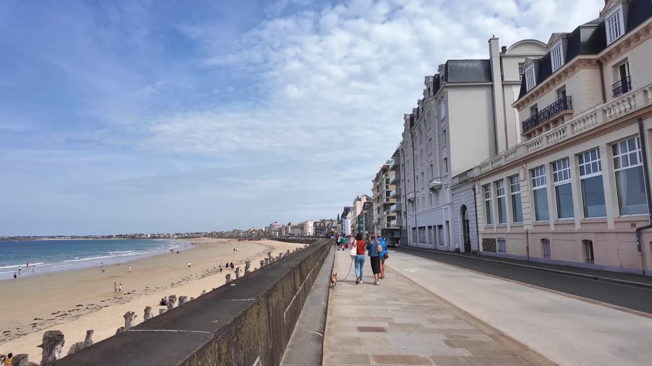 On the streets of Saint Malo, coastal promenade with people walking, sandy beach and row of houses. Brittany, France