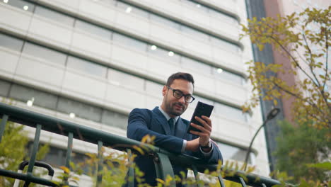 Un-Hombre-De-Negocios-Feliz-Se-Apoya-En-La-Barandilla-En-La-Calle-Del-Centro-De-La-Ciudad-Mirando-La-Pantalla-Del-Teléfono---ángulo-Bajo,-Cámara-Lenta