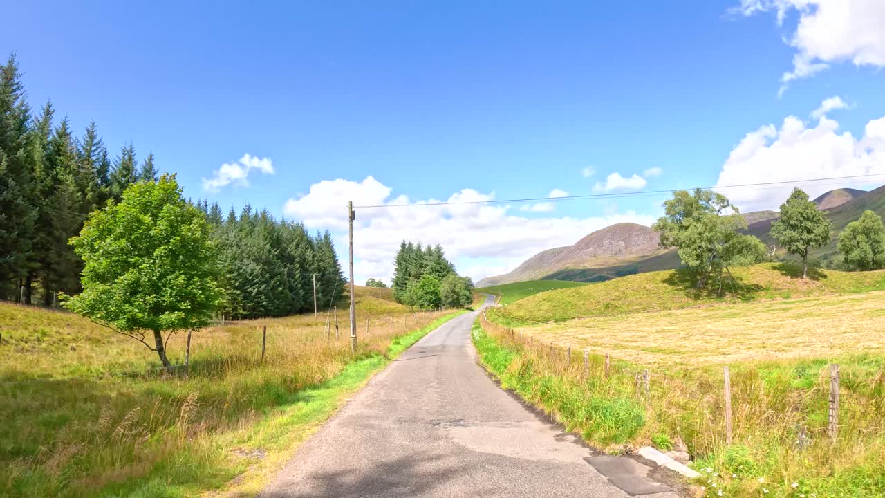 A vehicle travels a winding, tree-lined country road through grassy hills and farmland under bright daylight in Clova, Angus, Scotland. Smooth forward camera movement