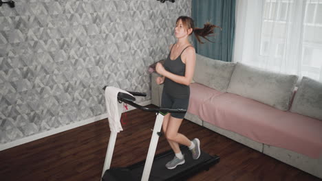 fitness lover running confidently on treadmill indoors, arms swinging rhythmically, hair tied in ponytail sways with movement, dressed in dark sportswear, towel draped over handle