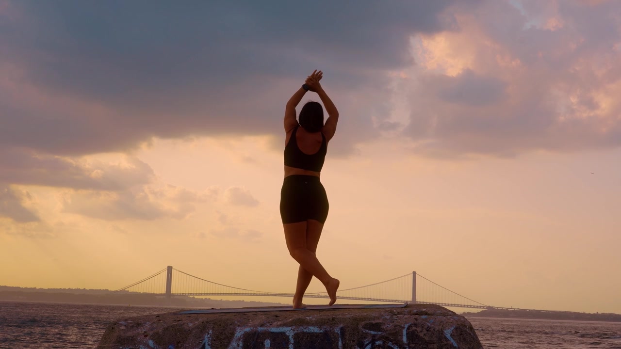Donna che esegue una posa yoga in piedi con le mani alzate al tramonto a New York. Vista posteriore di una figura femminile su un blocco di pietra vicino all'acqua con il ponte Verrazzano Narrows in lontananza