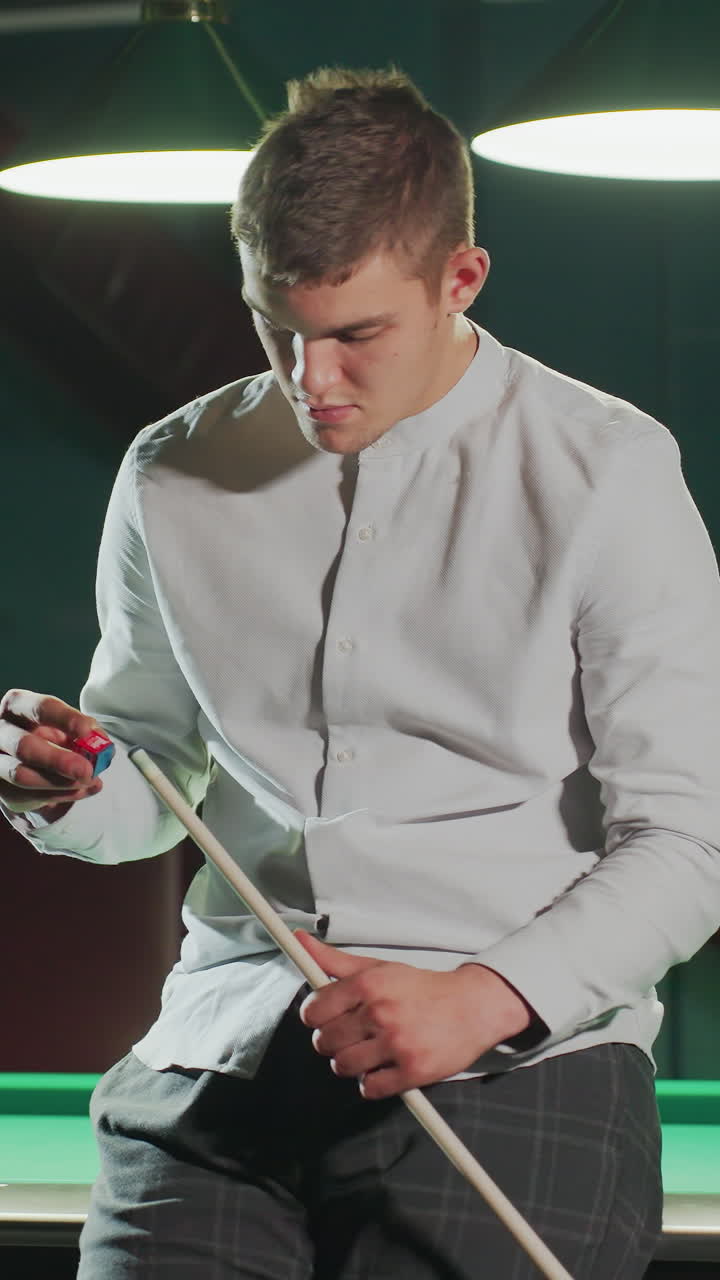 색 셔츠와 회색 바지를 입은 선수가 녹색 빌리어드 테이블에 앉아 큐 스틱을 들고 집중된 표정으로 은 개를 조심스럽게 적용합니다.