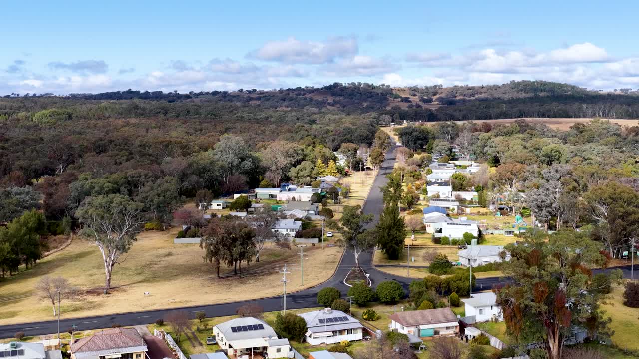 Drone footage glides above a small Australian town, revealing houses, tree-lined streets, and surrounding countryside under bright daylight with clear skies