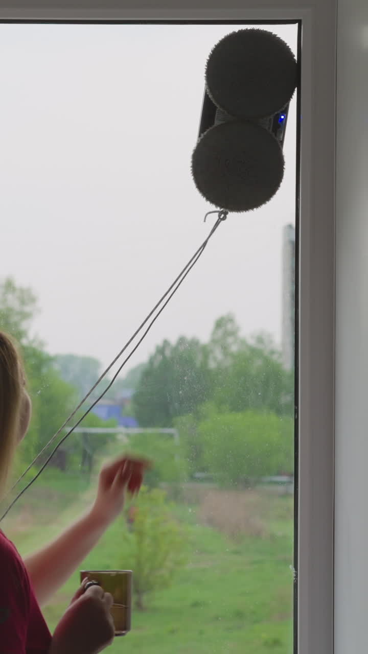 Long haired woman with tea mug taps on glass while contemporary automatic robot cleaner washes window at home backside view slow motion