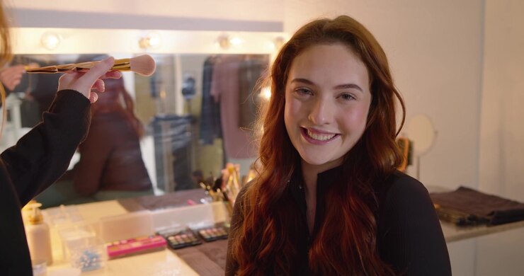 Makeup artist applying powder with large brush at vanity bulbs after client sits, female smiling