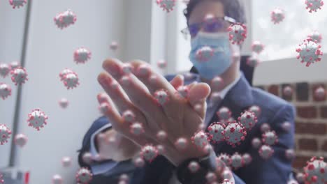 Animation-of-covid-19-cells-floating-over-man-wearing-face-mask,-sanitizing-his-hands-in-office