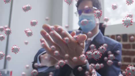 animation of covid 19 cells floating over man wearing face mask, sanitizing his hands in office