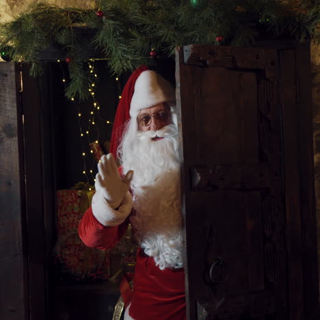 santa look out from the door and waving hand in white glove. wooden door and santa claus with white beard and eyeglasses making bye gesture. christmas time.