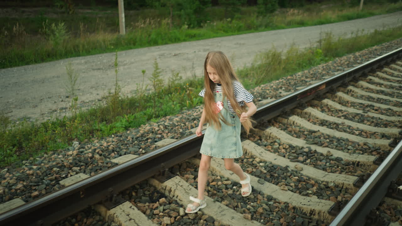 Young girl with long hair walking carefully along rail track holding grass strand in left hand, surrounded by rocky path and greenery, wearing striped shirt and denim dress under soft natural light