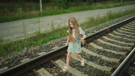 young girl with long hair walking carefully along rail track holding grass strand in left hand, surrounded by rocky path and greenery, wearing striped shirt and denim dress under soft natural light