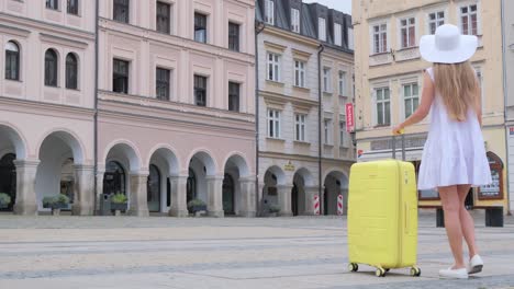 back view of a girl traveler walking and pulling a suitcase. a beautiful tourist girl in a white hat is traveling with a yellow suitcase. a woman enjoys a view of the city