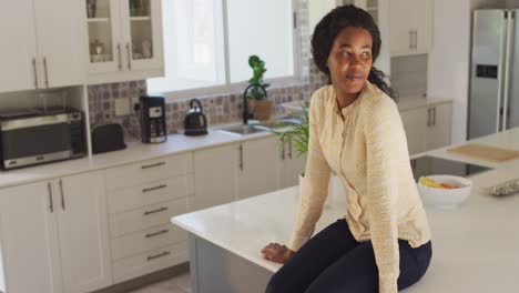 video of happy african american woman sitting on countertop in kitchen