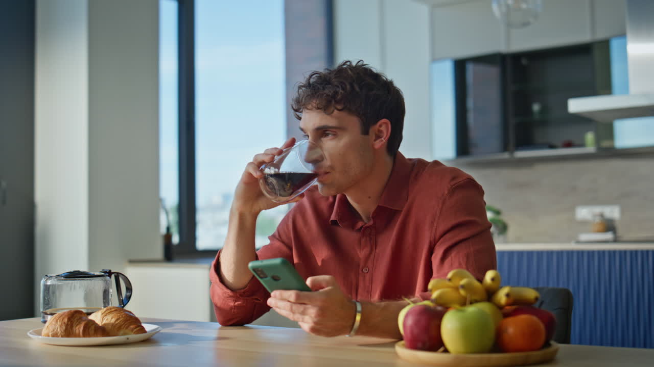 Relaxed man sipping coffee holding smartphone in kitchen closeup. Guy drinking