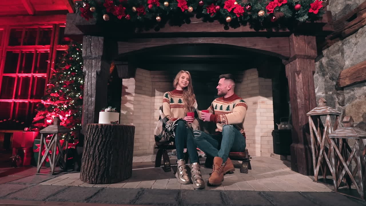 Couple in love spend time together at home. Young man and beautiful girl in knitted Christmas sweaters sitting at fireplace. Christmas background.