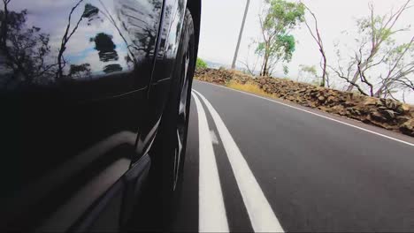 Punto-De-Vista-Del-Coche-Desde-La-Carretera-Mirando-Hacia-La-Rueda-Delantera-Derecha-Conduciendo-Por-Una-Carretera-Bituminosa-Bajando-Una-Colina-Hacia-La-Ciudad