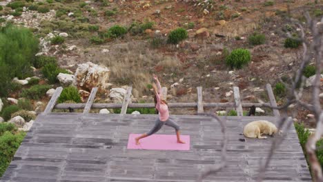 Caucasian-woman-practicing-yoga-outdoors,-stretching-standing-on-deck-in-rural-mountainside-setting