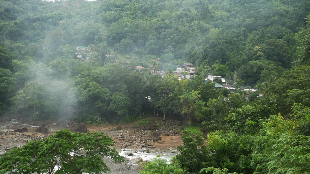Framed from a hillside angle, a small Philippine village nestles in dense forest beside a rocky stream, with rising mist adding depth to the lush, tranquil landscape