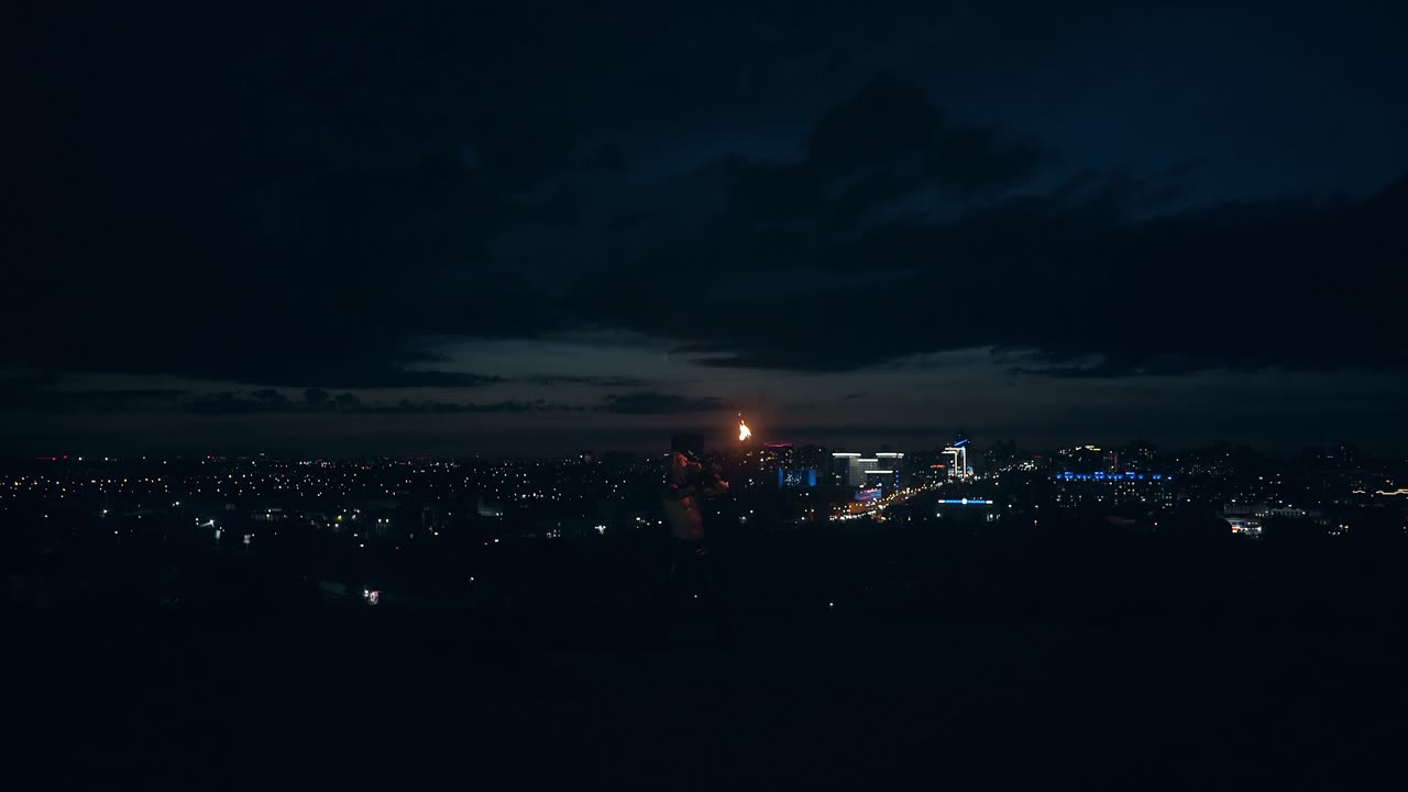 젊은 금발 남성이 불로 트릭을 하고 밤중에 불을  ⁇ 고 배경에는 도시의 스카이라인이 있습니다.