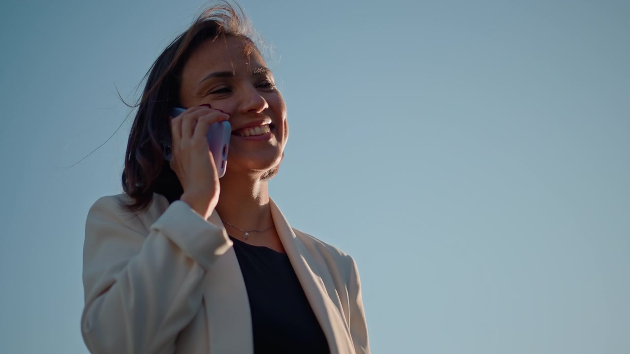 Confident young woman stands outdoors holding smartphone near ear while speaking, slight head tilt and bright smile conveying engaged upbeat mood beneath open blue sky in daylight, with woman, phone