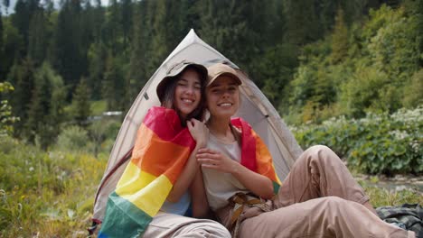 Porträt-Zweier-Lesbischer-Mädchen,-Die-Sich-In-Die-LGBT-Flagge-Gehüllt-Haben-Und-Neben-Einem-Zelt-Vor-Dem-Hintergrund-Eines-Grünen-Waldes-Sitzen.-Kampagne-Von-Menschen-Mit-Nicht-traditioneller-Sexueller-Orientierung