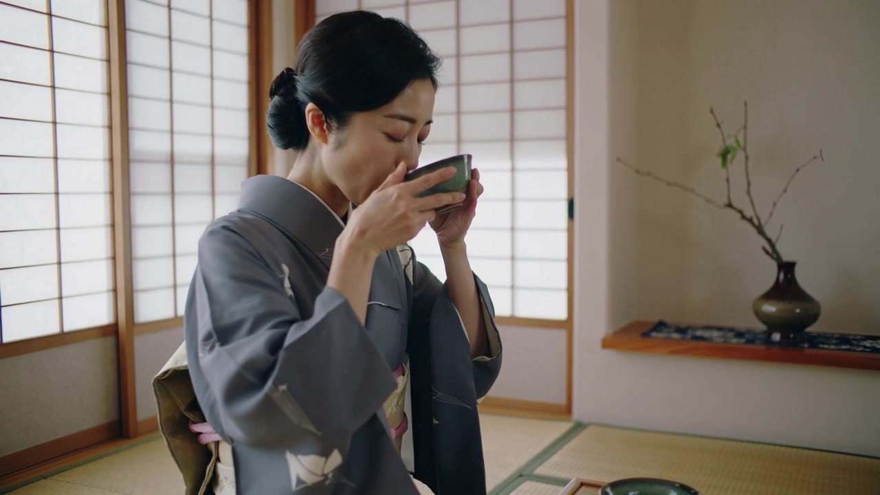 緑の茶碗を持ち、灰色の着物を着た女性が和室で茶碗を唇に近づけ、儀式を行っている。