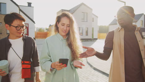 young multiethnic friends walking on street, using smartphone and talking