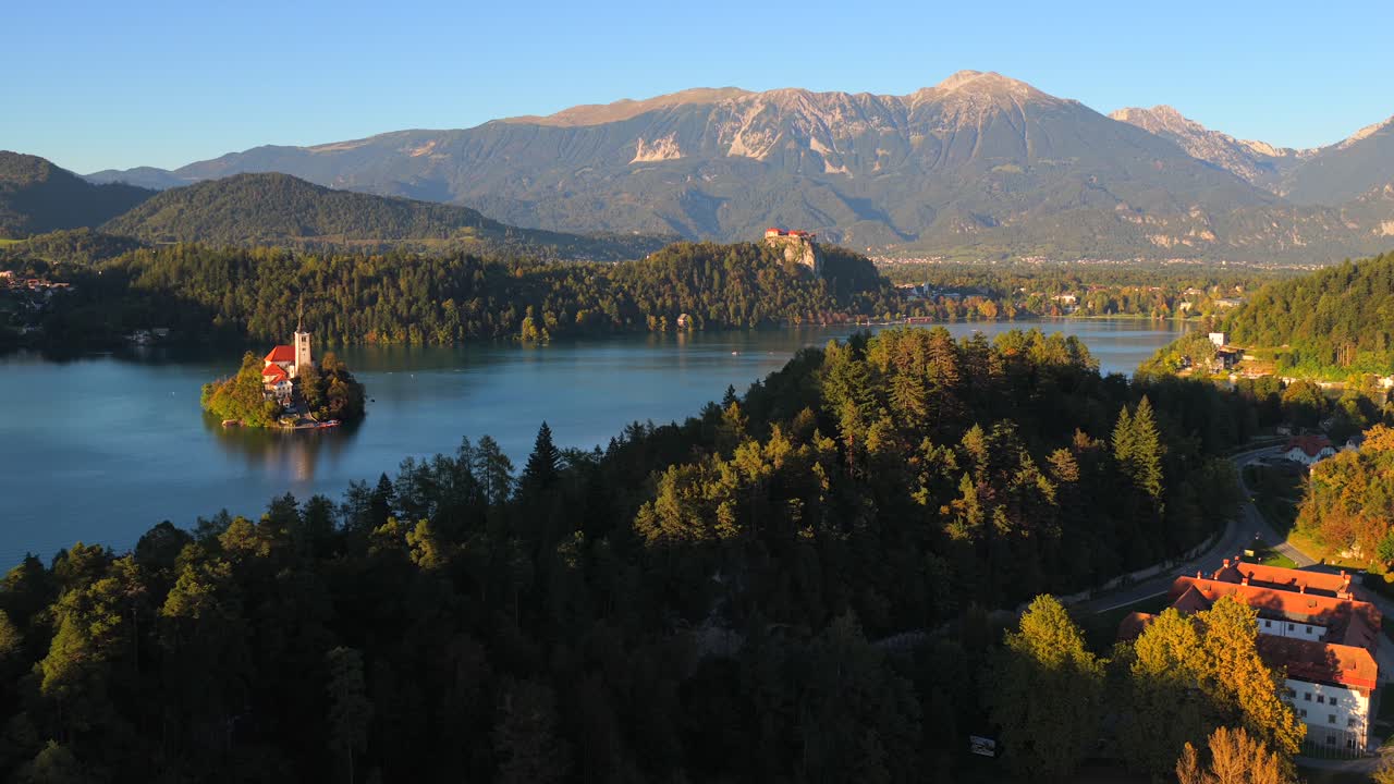 Ripresa aerea che mostra la chiesa sull'isola di Bled e il castello con acqua calma del lago e montagne nella tenue luce del giorno, stabilendo una luce dorata