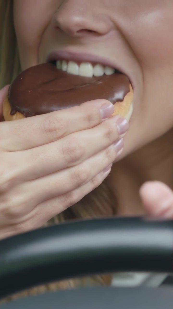 Vertical video: Picking up glazed donut, woman bringing bite to mouth then biting in car at wheel