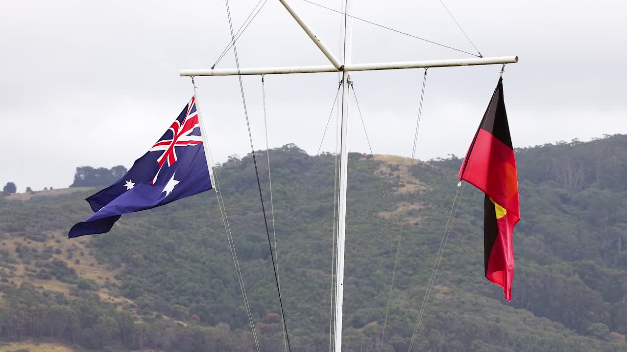 Australian and Aboriginal flags flutter against a hilly backdrop. Overcast lighting enhances the vibrant colors and dynamic movement