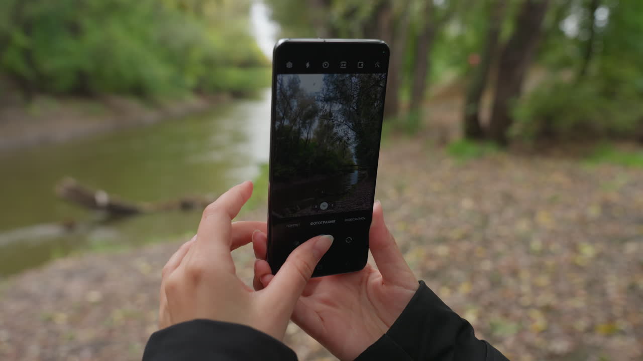 Pessoa caminhando perto de um rio, fotografando árvores e a margem com o celular, cabelo comprido e jaqueta preta visíveis, mãos enquadrando a foto na tela touchscreen, margem de seixos, ondulações na água calma e copa verde, nublado
