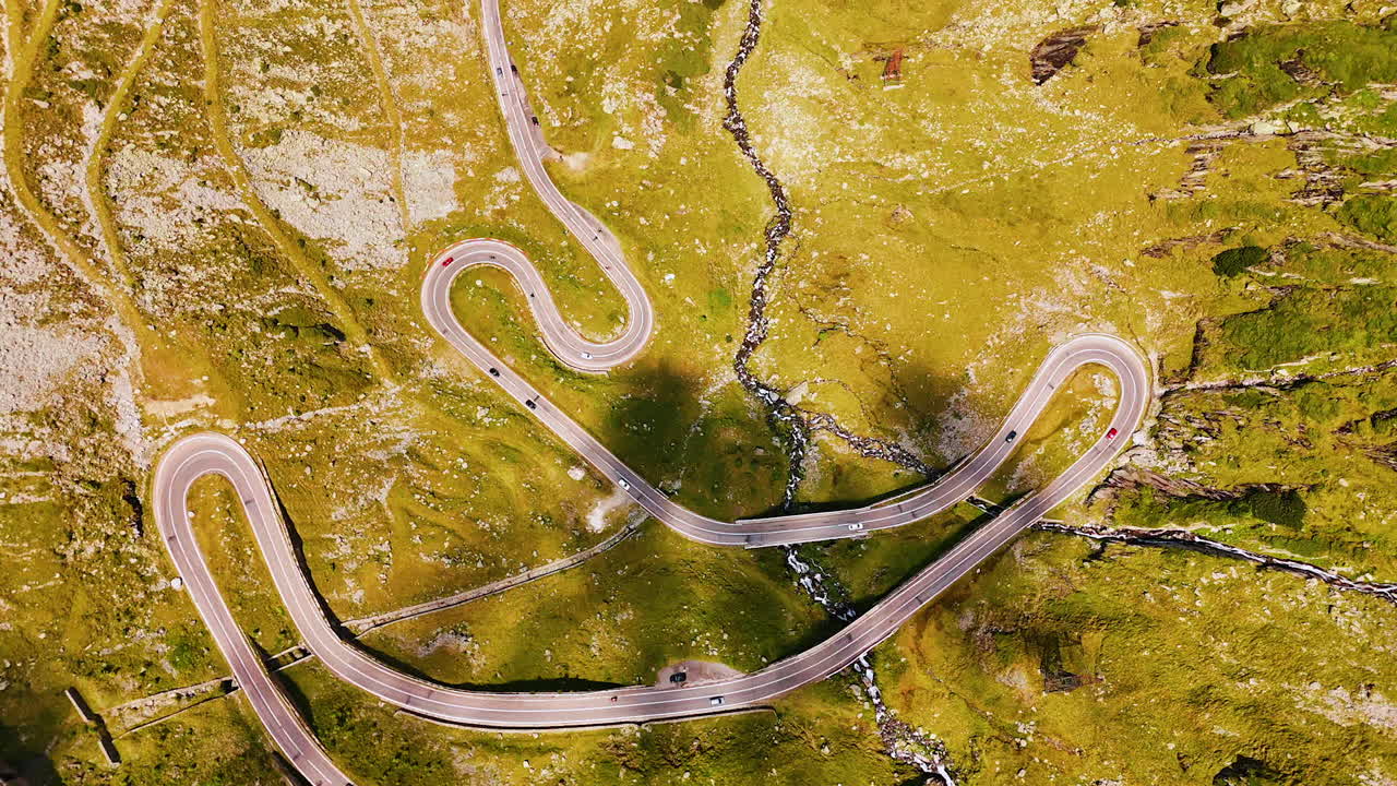Green slopes of the road crossed by the wavy road and narrow river. Footage over the Transfagarash highway in the Carpathian Mountains, Romania