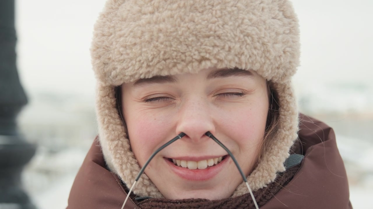 Smiling young woman in fleece-lined trapper hat and padded winter coat holds eyeglasses below chin, facing camera with relaxed pose, slight head tilt and soft overcast winter light