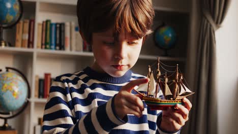 young boy in a striped sweater holds a model ship, his imagination taking him on voyages across the globe, inspired by the globes and books on the shelf behind him