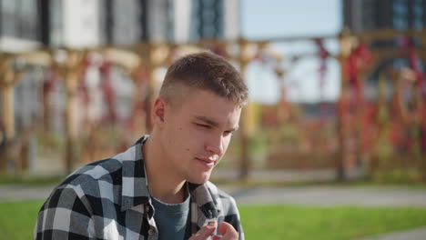 calm man wearing checkered shirt takes red pill medicine outdoors in sunny weather with focused expression and blurred background featuring tall buildings and wooden pergola covered in red vines