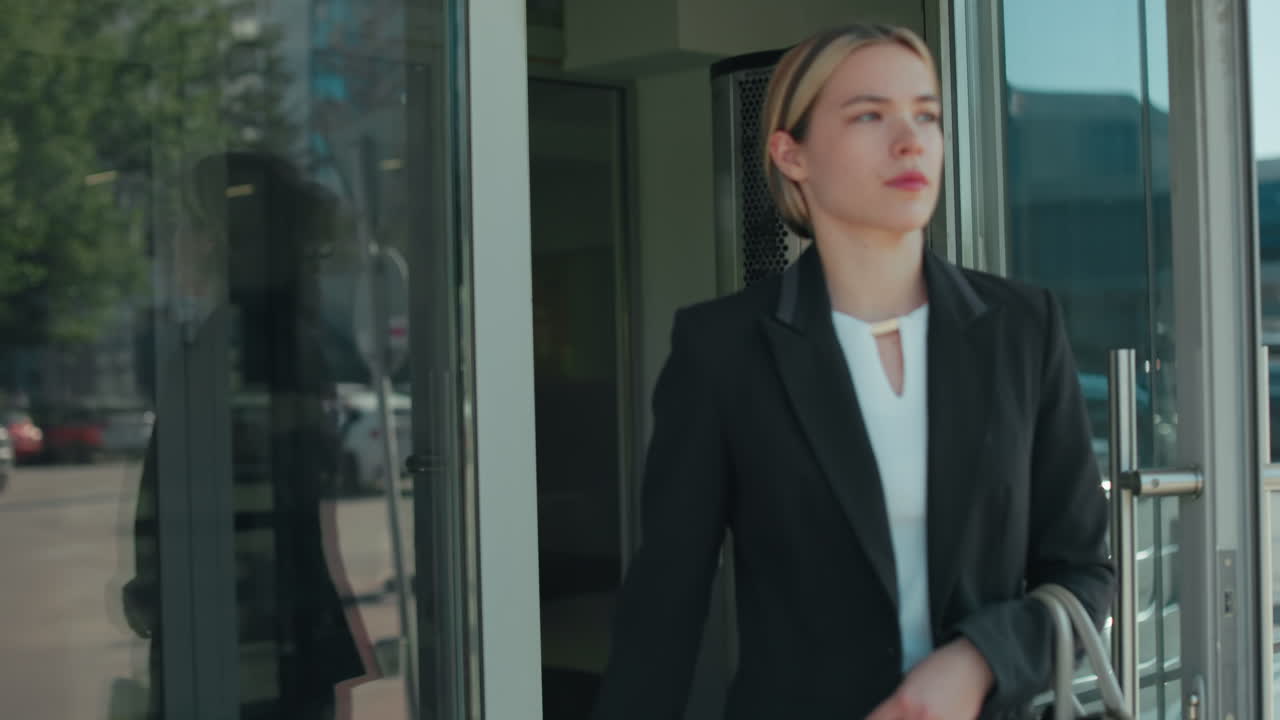 Confident woman walks out of door holding handbag with glass reflecting passing car and parked vehicles in urban setting, focus on motion, modern lifestyle, professional attire, and city environment