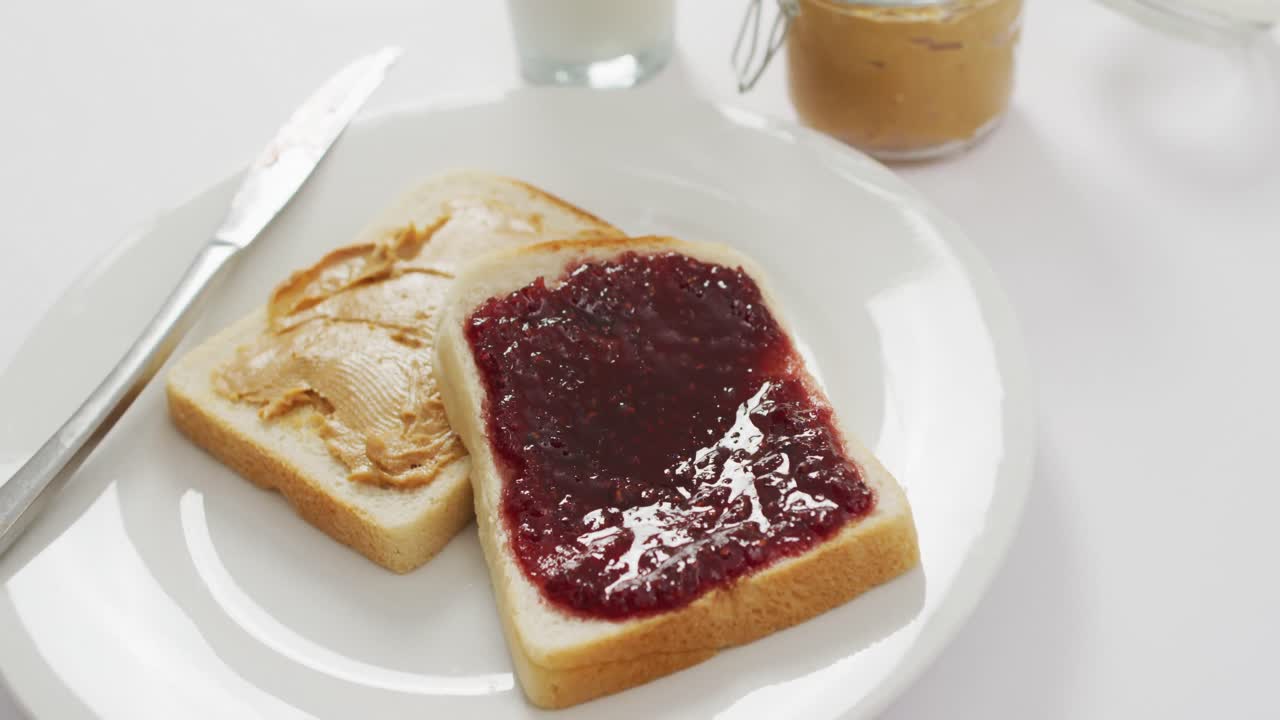 Närbild av en smörgås med jordnötssmör och sylt på en tallrik på en vit yta.