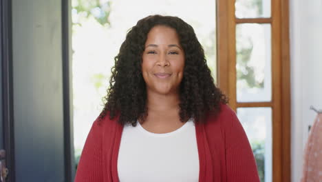 Happy-biracial-woman-with-long-curly-hair-standing-and-smiling-in-sunny-house