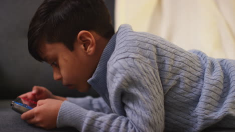 close up of young boy lying on sofa at home playing game or streaming onto handheld gaming device